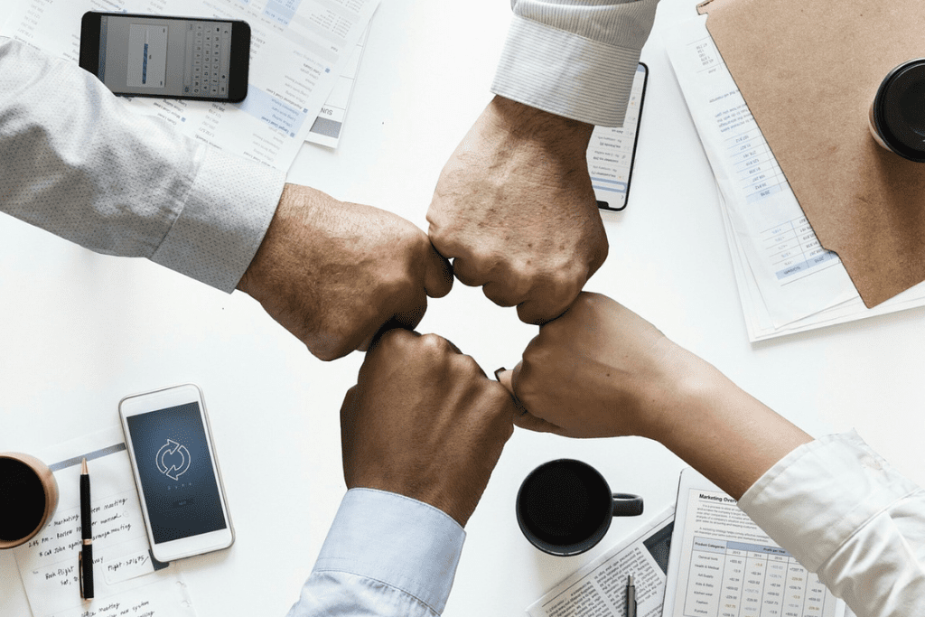 Four shirted hands clutching to each other by wrists over a desk with documents and smartphones on it.