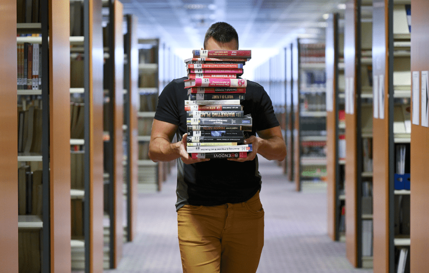 Young male person among bookshelves holding a pile of books hiding his face.