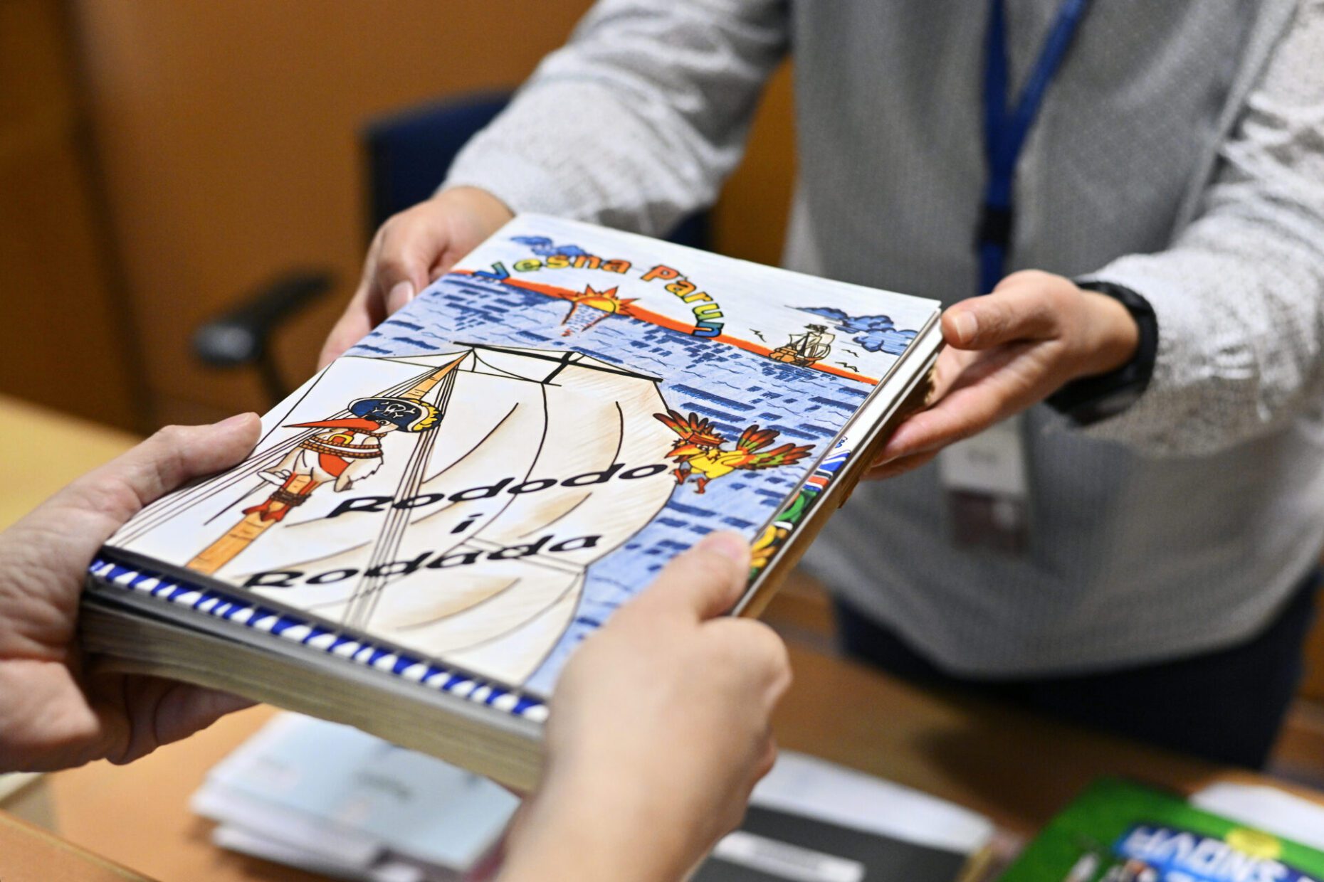 A library user handing a book with a colourful cover to a librarian, with only the user’s and librarian’s hands being visible.
